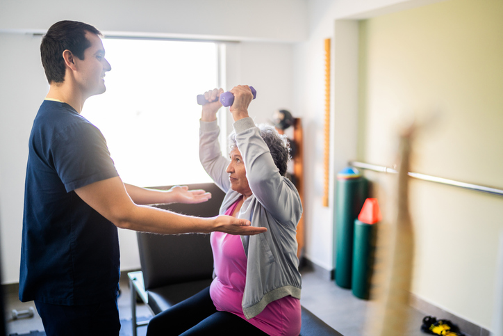 Mid adult physical therapist helping patient during exercise with dumbbell