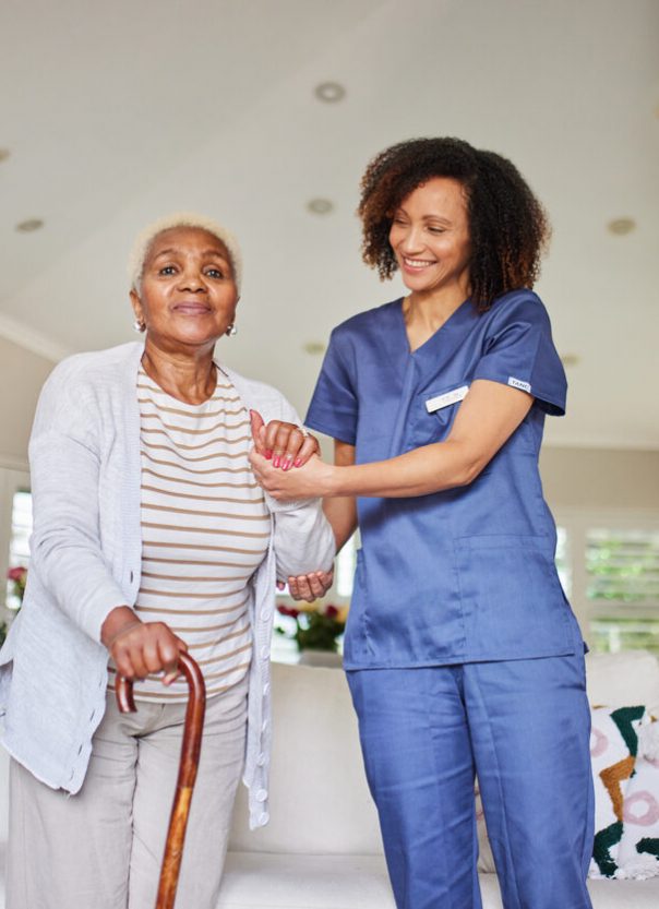 A kind caregiver in blue scrubs gently supports an elderly woman with a cane. They share a warm smile in a bright, comfortable living room setting.