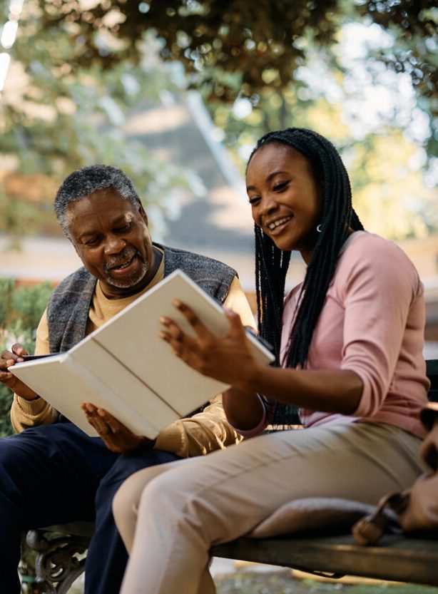 Happy black senior and his daughter looking at family photo album in the park at nursing home.