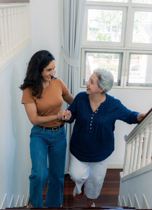 A beautiful mixed-race mid-adult woman holding and assisting her joyful retired senior mother stepping up the stairs in their house