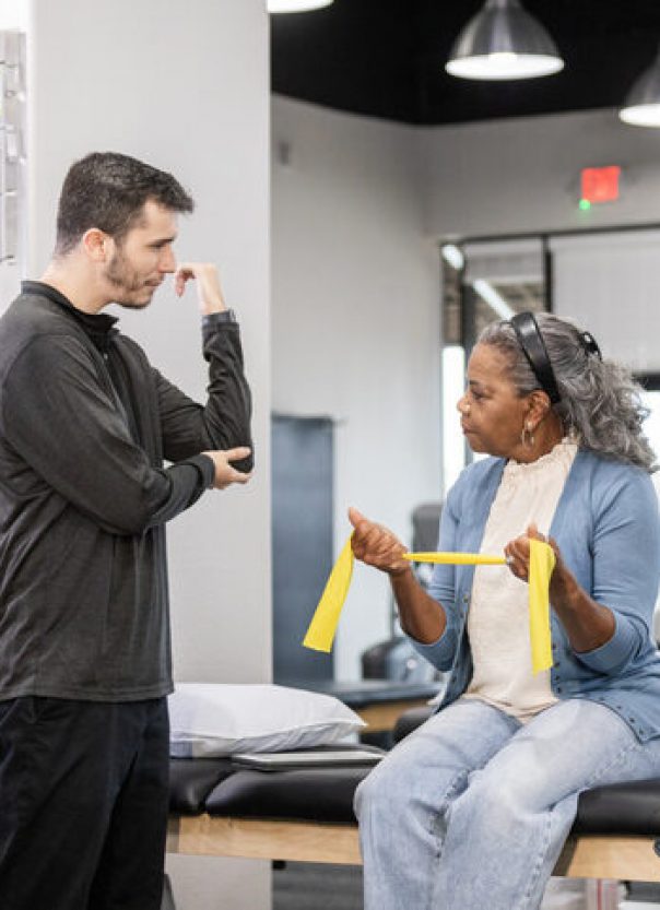The male occupational therapist gestures towards his elbow while he talks to the mature female patient.