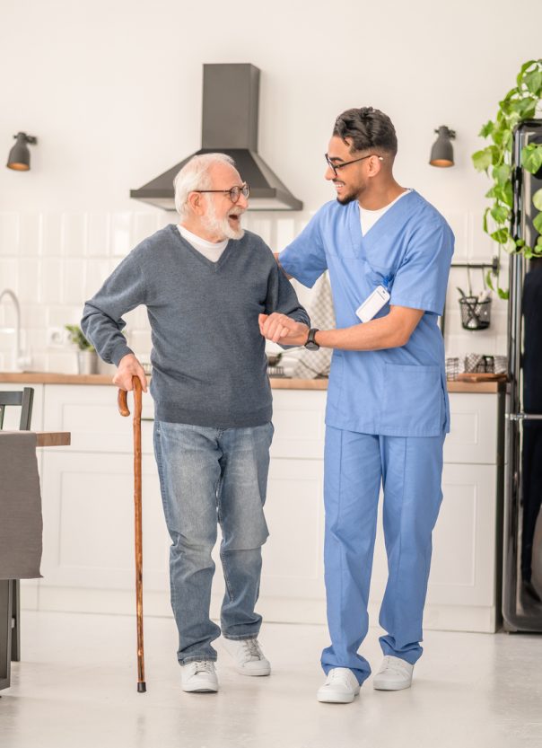 Full-length portrait of a cheerful aged man walking with a cane supported by a medical worker