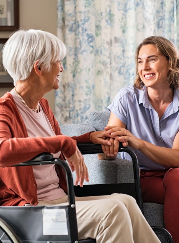 Mature woman comforting senior mom sitting on wheelchair at nursing home. Cheerful woman talking to old disabled mother in wheelchair at elder care centre. Loving caregiver taking care of elderly woman at home.