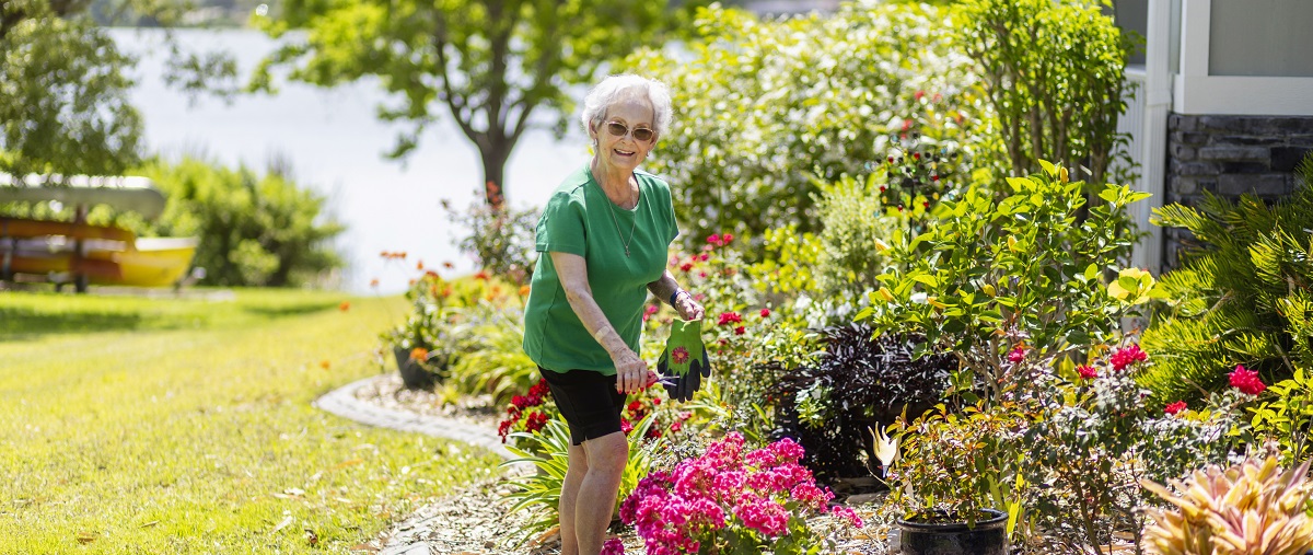 senior woman stands in her yard at Westminster Lakeland trimming flowers in a large flower bed