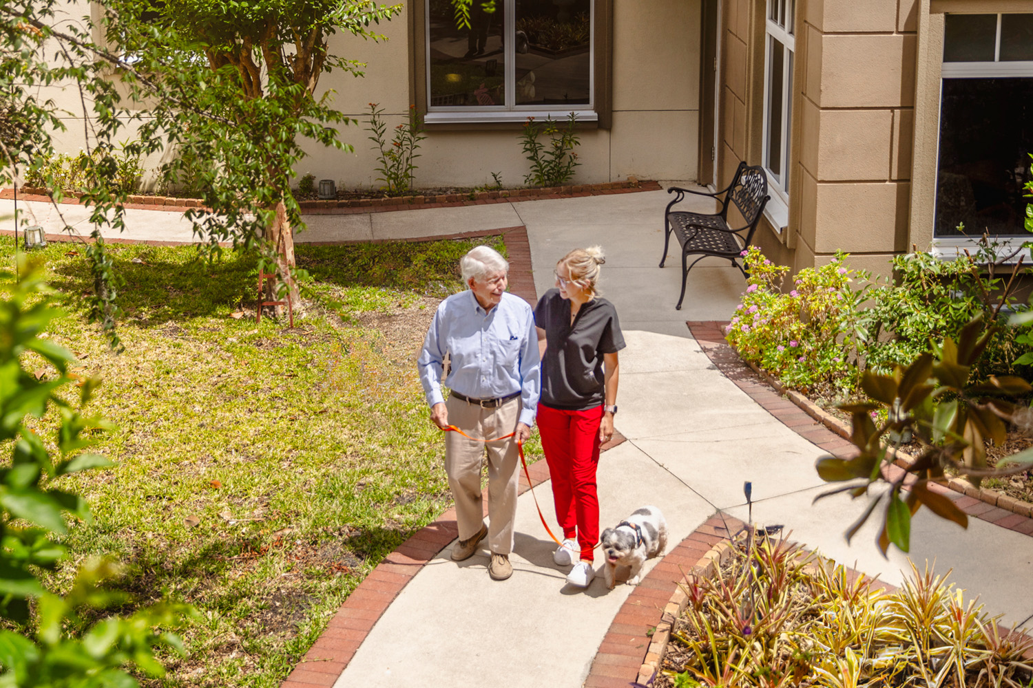 A senior man and a caregiver, a young woman, walk on a path outdoors with his dog.
