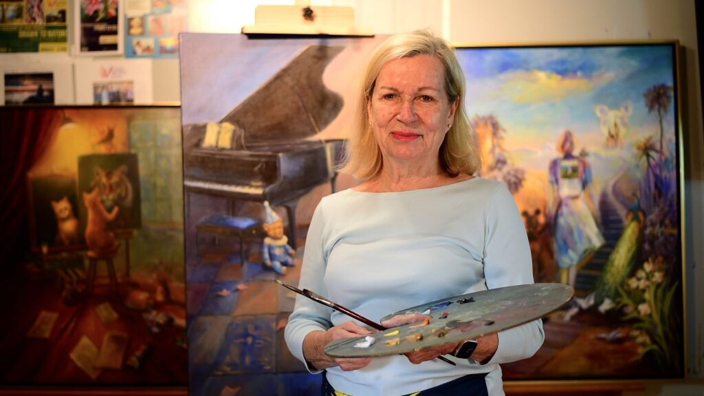 Artist Beatrice Athanas smiles in front of her artwork with a palette in her hand
