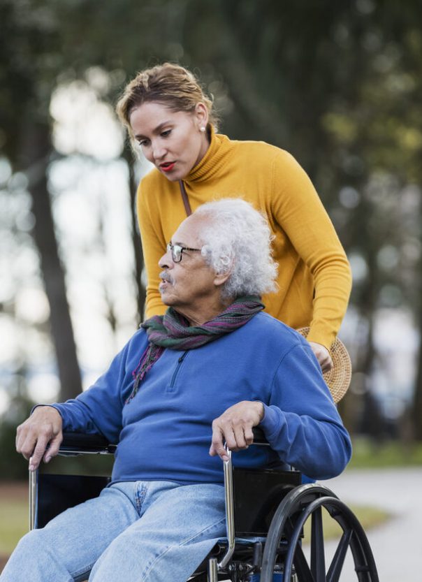 A senior Hispanic man in his 80s sitting in a wheelchair, taking a walk in the park with his adult daughter, a mid adult woman in her 30s.