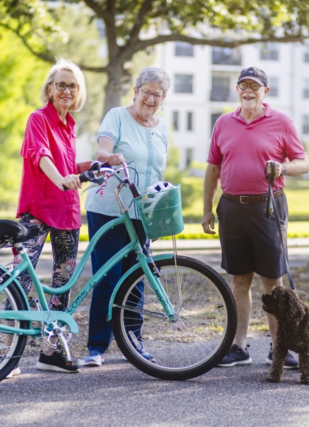 Seniors standing outside, one is on a bike, one has a bike, the other has a dog