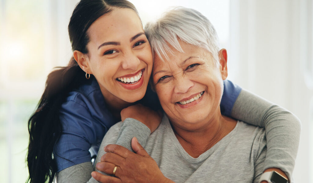 A caregiver and a senior woman embrace with big smiles