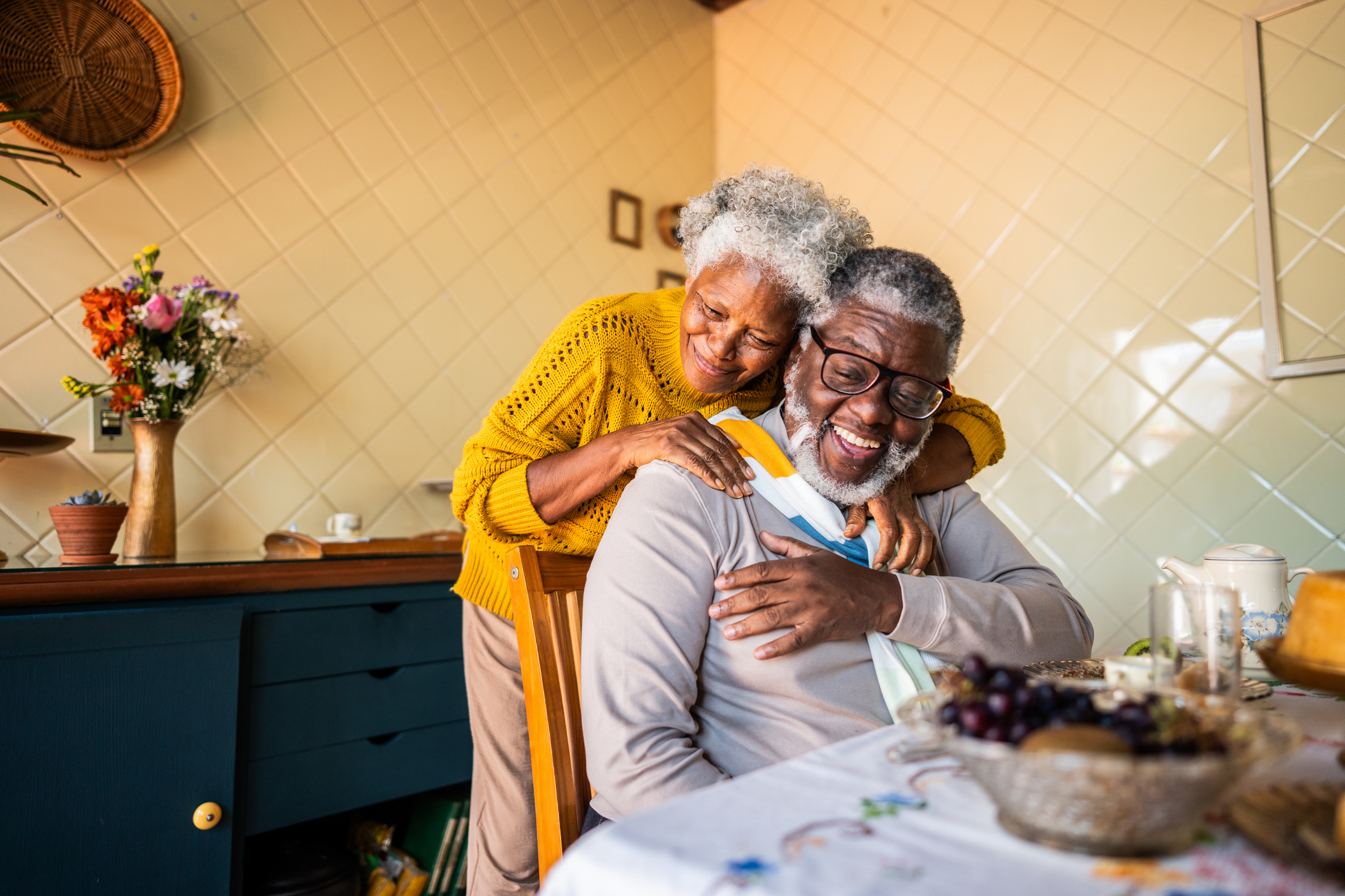 Senior woman hugs senior man who is sitting in a chair